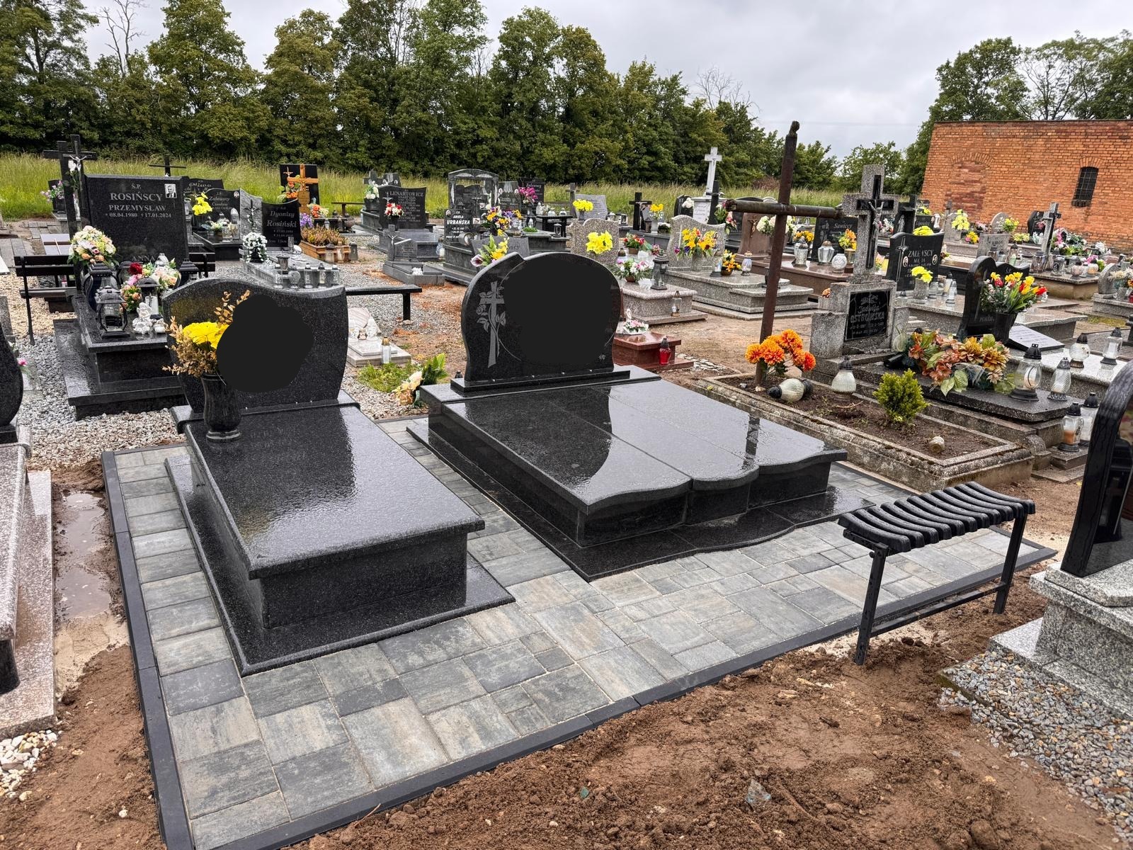 Dwa nowe, czarne nagrobki z granitu na cmentarzu, otoczone świeżo ułożoną kostką brukową. W tle inne groby i zieleń. Widok na nagrobki pod lekkim kątem.
