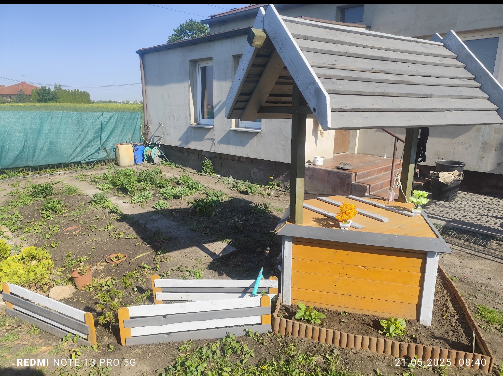 Ujęcie studni ogrodowej z szarym dachem i żółtą obudową, otoczonej rabatą z niskim płotkiem, na tle budynku z ocieploną elewacją i zielonej siatki.