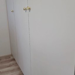 Kussa Meble - Minimalistyczna zabudowa meblowa z jasnymi, matowymi frontami i subtelnymi, okrągłymi, złotymi gałkami, na tle drewnopodobnej podłogi.