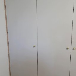 Kussa Meble - Szara szafa z czterema gładkimi drzwiami, złotymi gałkami i drewnianymi obramowaniami, zabudowana na całej wysokości ściany.