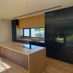 Kussa Meble - Nowoczesna kuchnia z jasną drewnianą wyspą ze żaluzjowym frontem i płytą indukcyjną, czarnymi szafkami w zabudowie z piekarnikiem (częściowo w folii ochronnej) i górnymi szafkami z jasnego drewna.