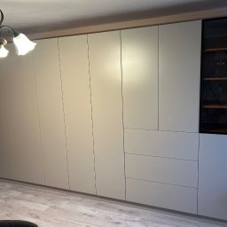 Kussa Meble - Minimalistyczna zabudowa meblowa w kolorze greige, z matowymi frontami bez uchwytów, przeszkloną witryną z drewnianymi półkami i trzema szufladami, oświetlona żyrandolem.