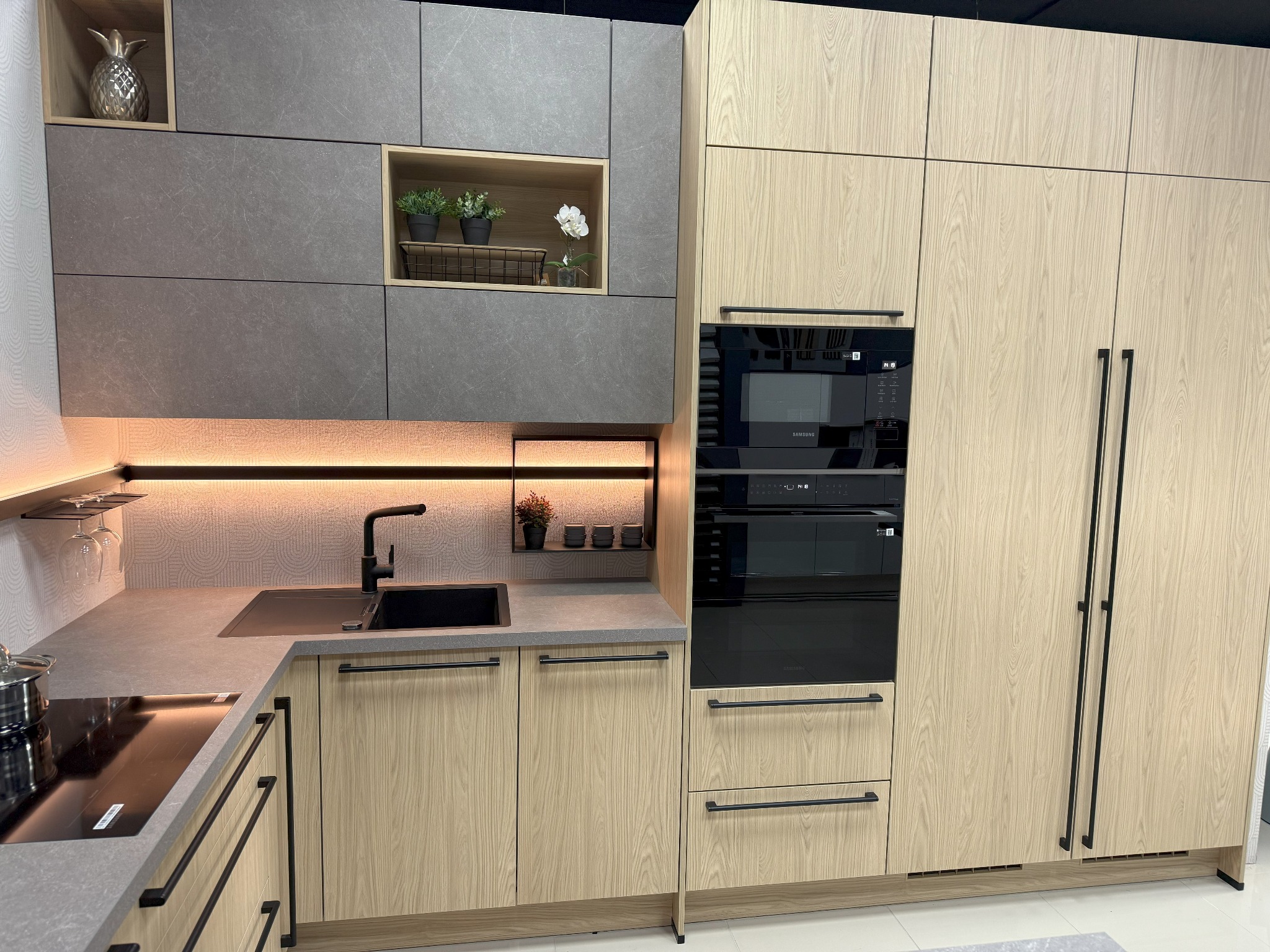 Nowoczesna, jasna kuchnia na wymiar z drewnopodobnymi frontami, czarną armaturą i sprzętem AGD. Na blacie widoczny garnek i płyta indukcyjna. Dekoracje w półkach.