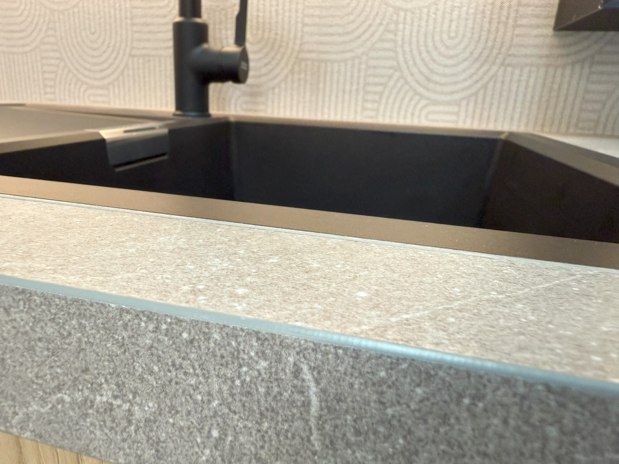 Minimalistyczny, czarny zlew wpuszczony w blat z szarego laminatu. Bateria w kolorze czarnym. W tle wzorzysta tapeta.