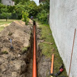 OLMAX Jakub Olejnik - Wkopana pomarańczowa rura w wąskim wykopie na trawiastym terenie, narzędzia pracy obok. Osoba pracuje w oddali, domy i drzewa w tle.