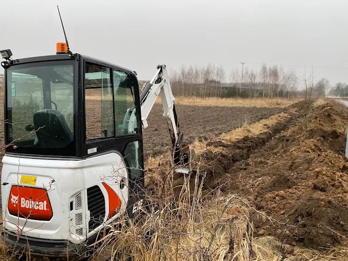 Mini koparka Bobcat wykonuje wykop rowu w ziemi, widok z boku, krajobraz polny w tle, pochmurny dzień.