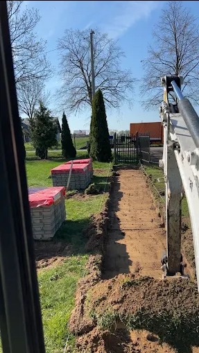 Wąski wykop pod trawnikiem, wyznaczony sznurkiem, z widocznymi kostkami brukowymi w tle i ramieniem koparki po prawej, wykonany w słoneczny dzień.