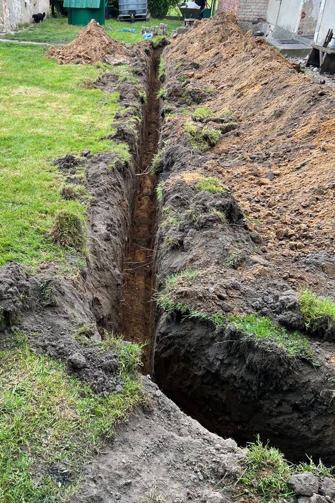 Długi, wąski wykop w ziemi z równo ułożoną darnią po bokach, przygotowany pod instalacje. W tle widoczny sprzęt budowlany i częściowo budynek.