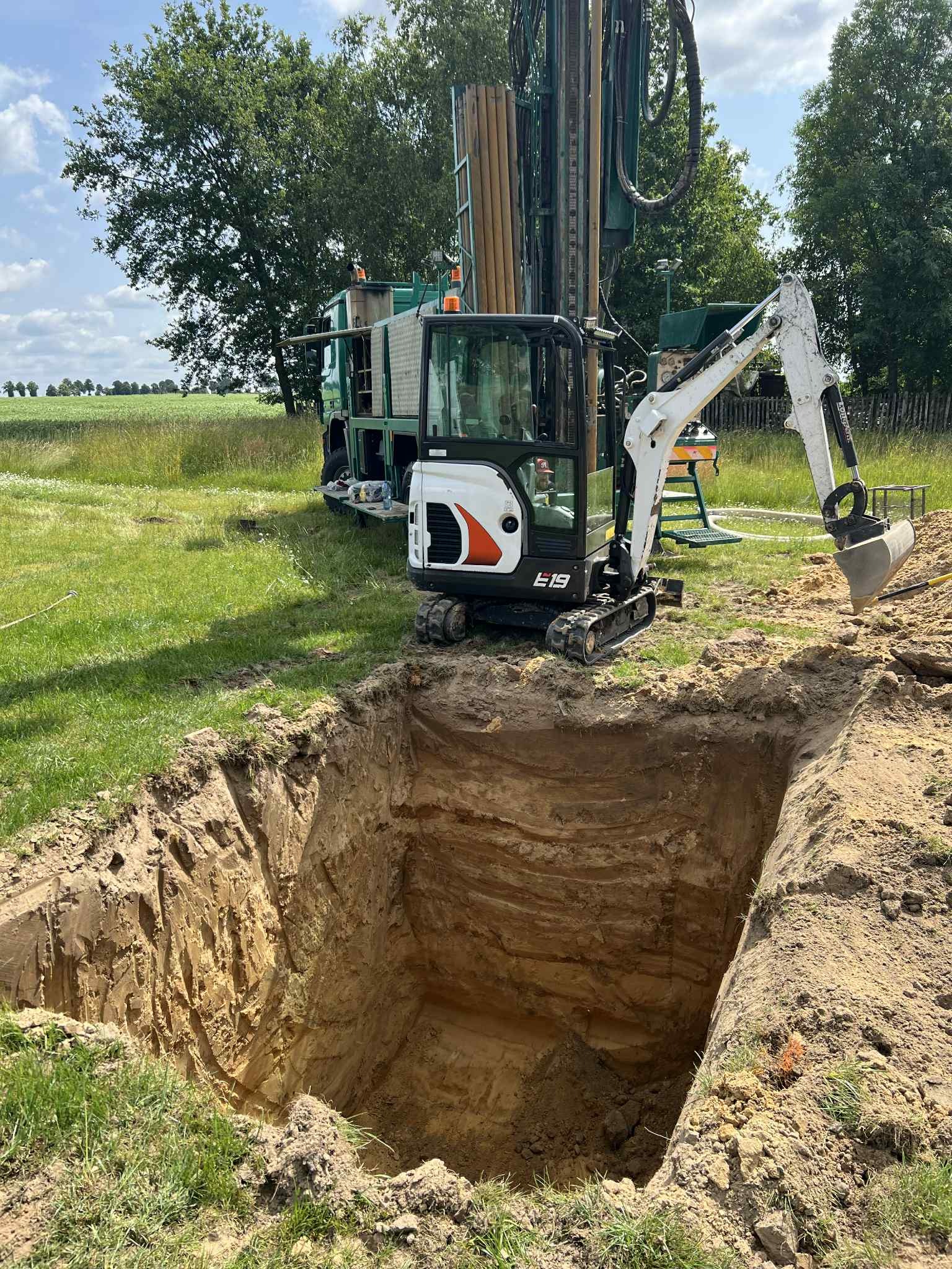 Głęboki wykop w ziemi, z boku stoi mała koparka E19 i wiertnica na tle zielonego pola i nieba. Przekrój ziemi z warstwami piasku i gliny.