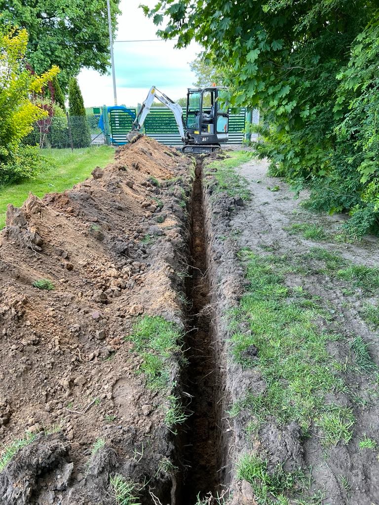 Długi, wąski wykop w ziemi, wykonany przez małą koparkę na tle zielonego trawnika i ogrodzenia z metalowych sztachet. Prace ziemne w toku.