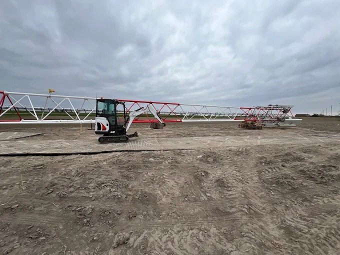 Mini koparka Bobcat obok długiej, poziomej konstrukcji kratownicowej, częściowo pomalowanej na biało i czerwono, na przygotowanym terenie pod budowę.