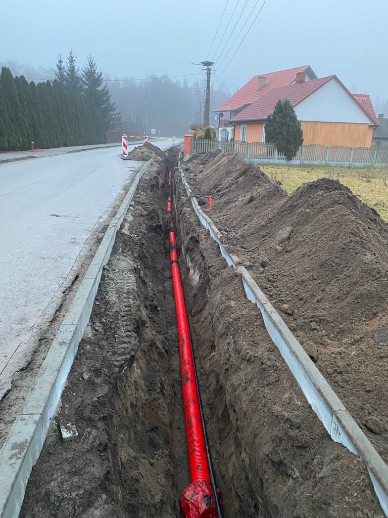 Czerwona rura w wykopie wzdłuż chodnika, widok z góry. Prace ziemne na tle zabudowań i drzew w pochmurny dzień. Etap instalacji podziemnej.