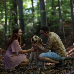 Mateusz Szewczyk - Para w lesie głaszcze golden retrievera. Kobieta w sukience i mężczyzna w koszuli w kratę kucają obok psa na tle drzew i opadłych liści.