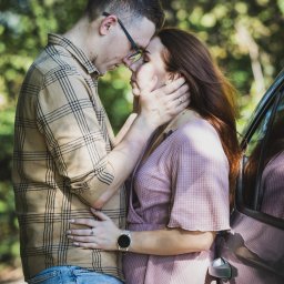 Mateusz Szewczyk - Zakochana para oparta o samochód, dotykająca się czołami na tle zieleni. Mężczyzna w kraciastej koszuli i jeansach, kobieta w różowej sukience. Romantyczna sesja.