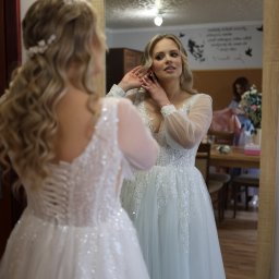 Mateusz Szewczyk - Panna młoda w białej sukni z koronkowym gorsetem i tiulowym dołem, poprawia kolczyk przed lustrem. W tle widoczna osoba i kwiaty.