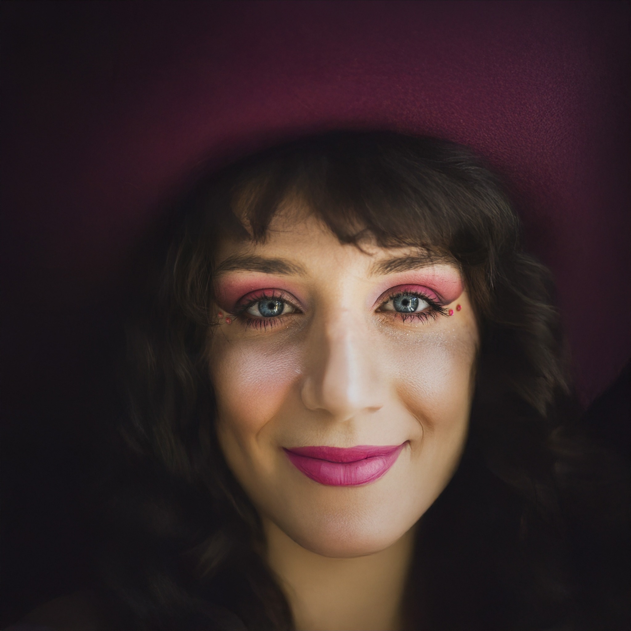 Portret kobiety z artystycznym makijażem w odcieniach różu i burgundu, z uwydatnionymi niebieskimi oczami i ciemnymi, kręconymi włosami, na ciemnym tle.