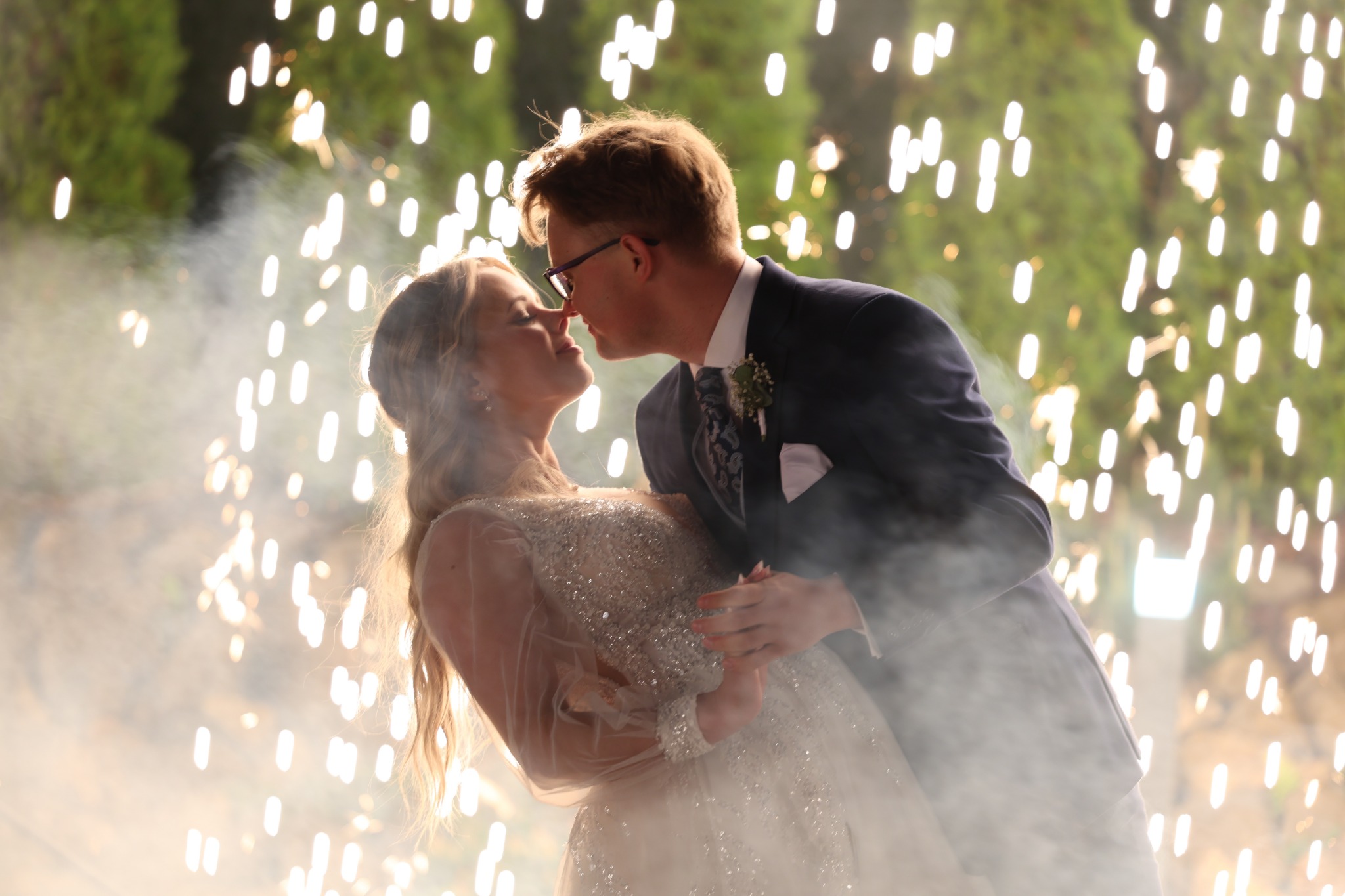 Para młoda w objęciach, zbliżająca się do pocałunku na tle fontanny iskier. Panna młoda w zdobionej sukni, pan młody w garniturze z butonierką. Romantyczna atmosfera.