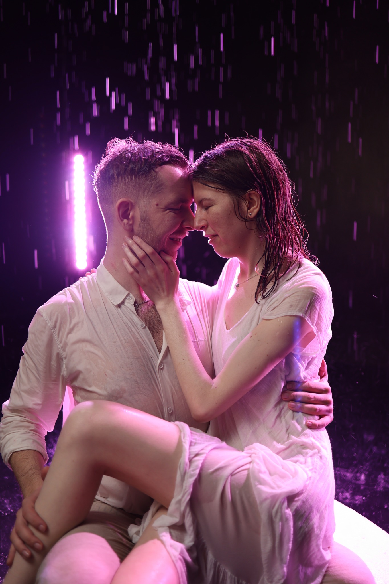 Para w mokrych ubraniach, obejmująca się w deszczu, oświetlona fioletowym światłem, tworząca intymną i romantyczną scenę. Ujęcie z bliska, skupione na emocjach.
