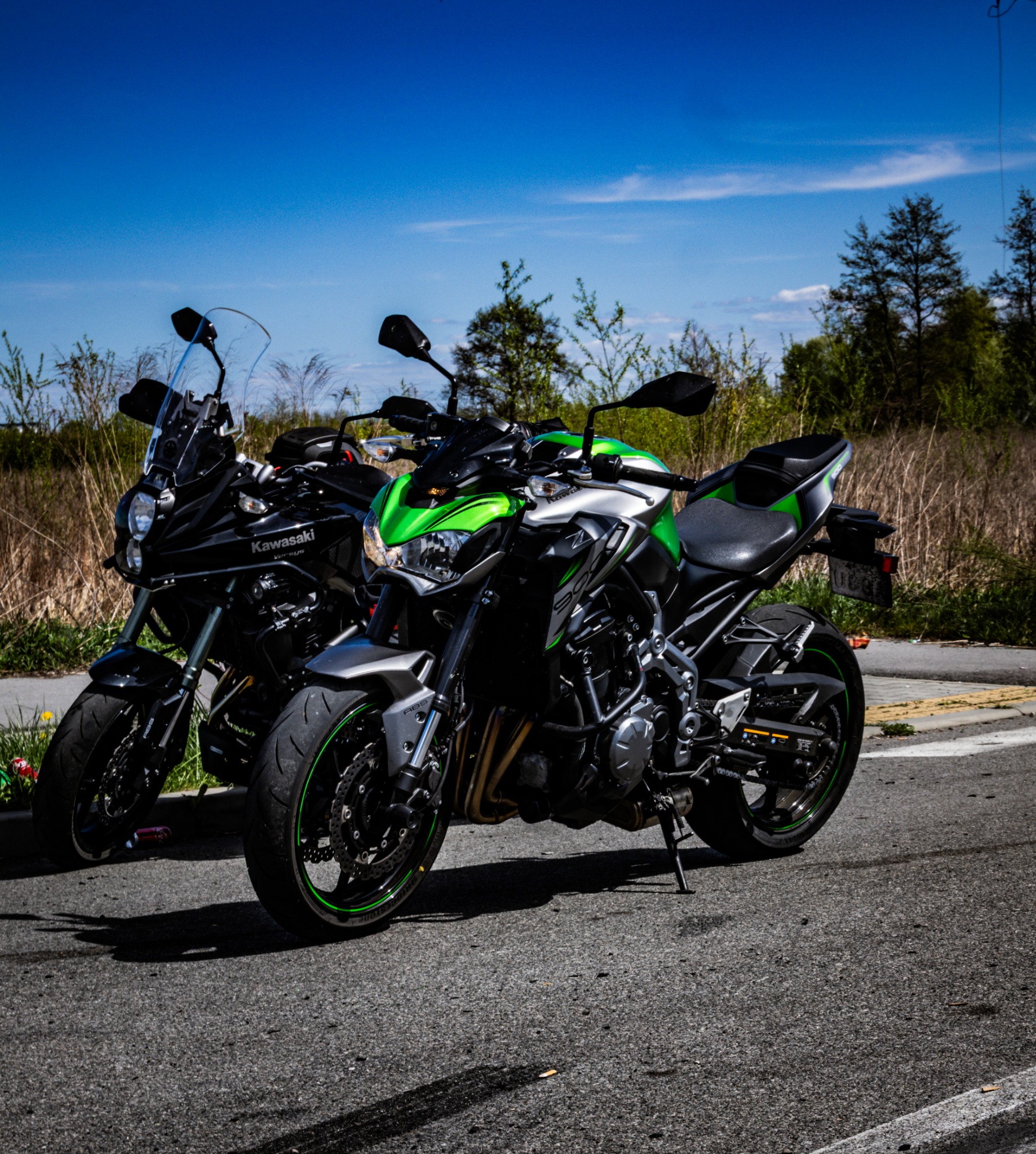 Dwa motocykle Kawasaki: czarny Versys i srebrno-zielony Z900, zaparkowane obok siebie na asfalcie, z zielenią i błękitnym niebem w tle.