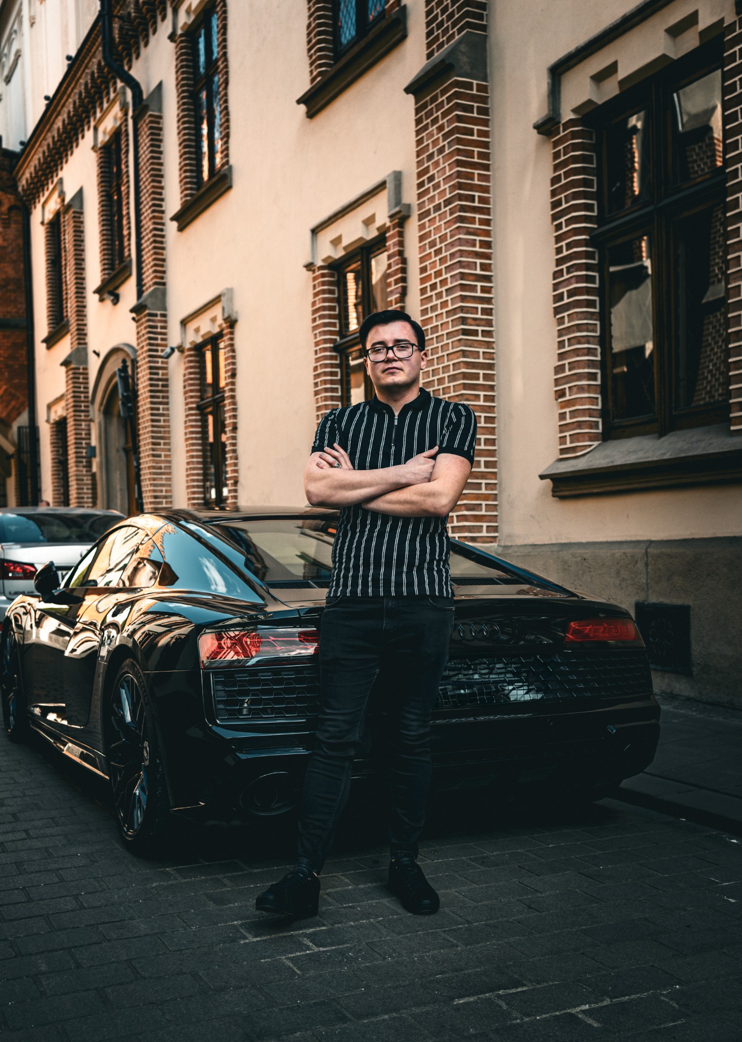 Mężczyzna w okularach, z założonymi rękoma, stoi przed czarnym Audi R8 na tle kamienicy z cegły w Krakowie. Ujęcie z dołu podkreśla auto i architekturę.