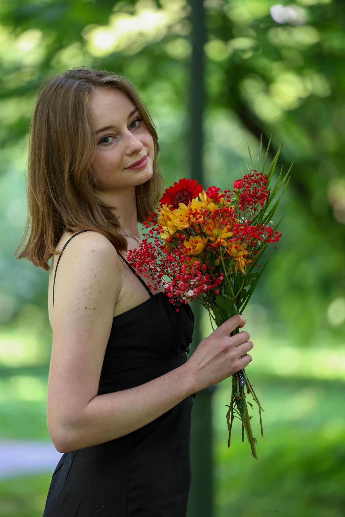 Portret młodej kobiety w czarnej sukience trzymającej bukiet czerwono-żółtych kwiatów na tle zieleni. Naturalne światło, delikatny uśmiech, swobodna poza.