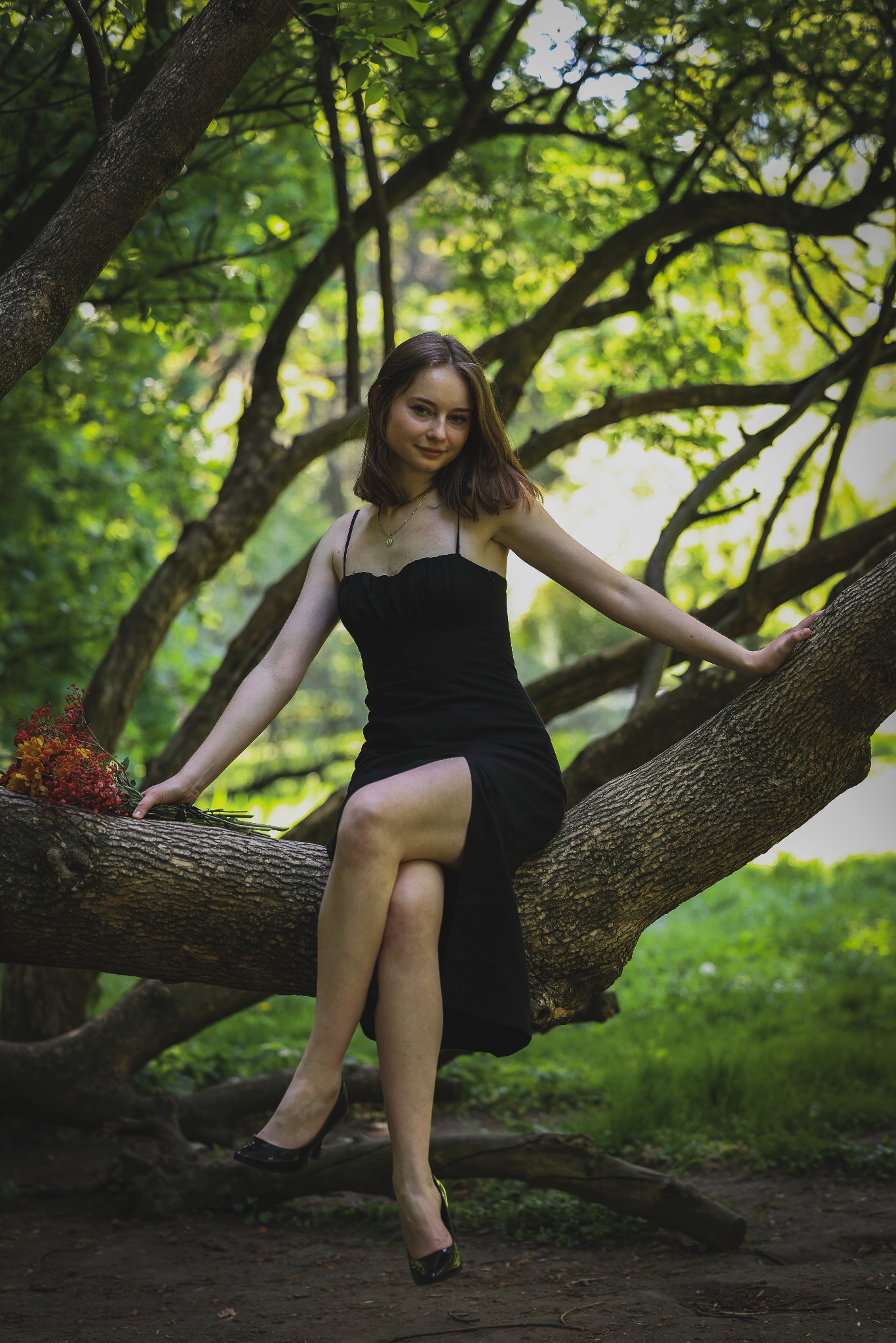 Kobieta w czarnej sukience siedzi na gałęzi drzewa w lesie, trzymając bukiet kwiatów. Uśmiechnięta, patrzy w obiektyw. Naturalne światło, zielone tło.