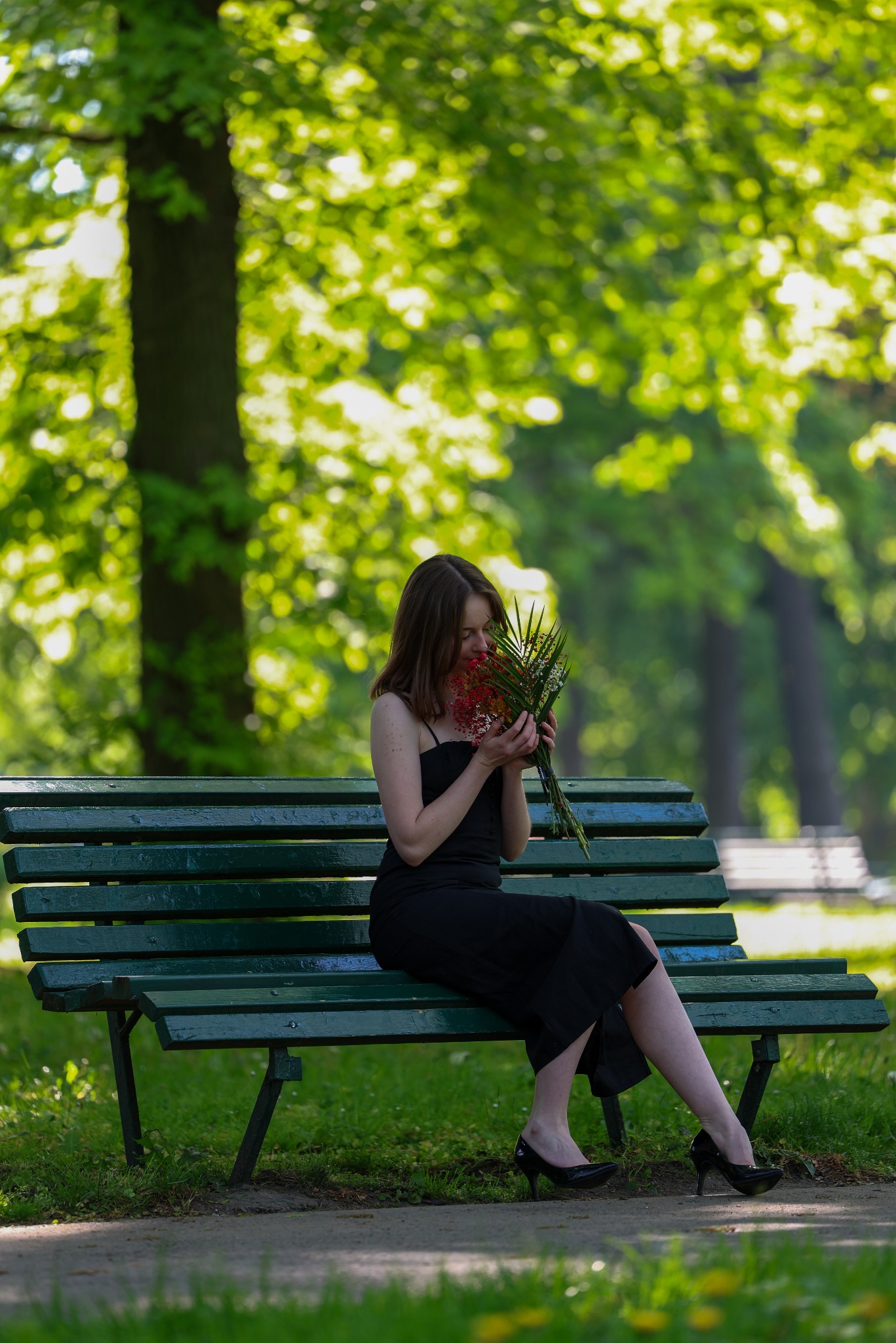 Kobieta w czarnej sukience siedzi na zielonej ławce w parku, wąchając bukiet czerwonych kwiatów. Słoneczny dzień, zielone drzewa w tle, eleganckie czarne buty.
