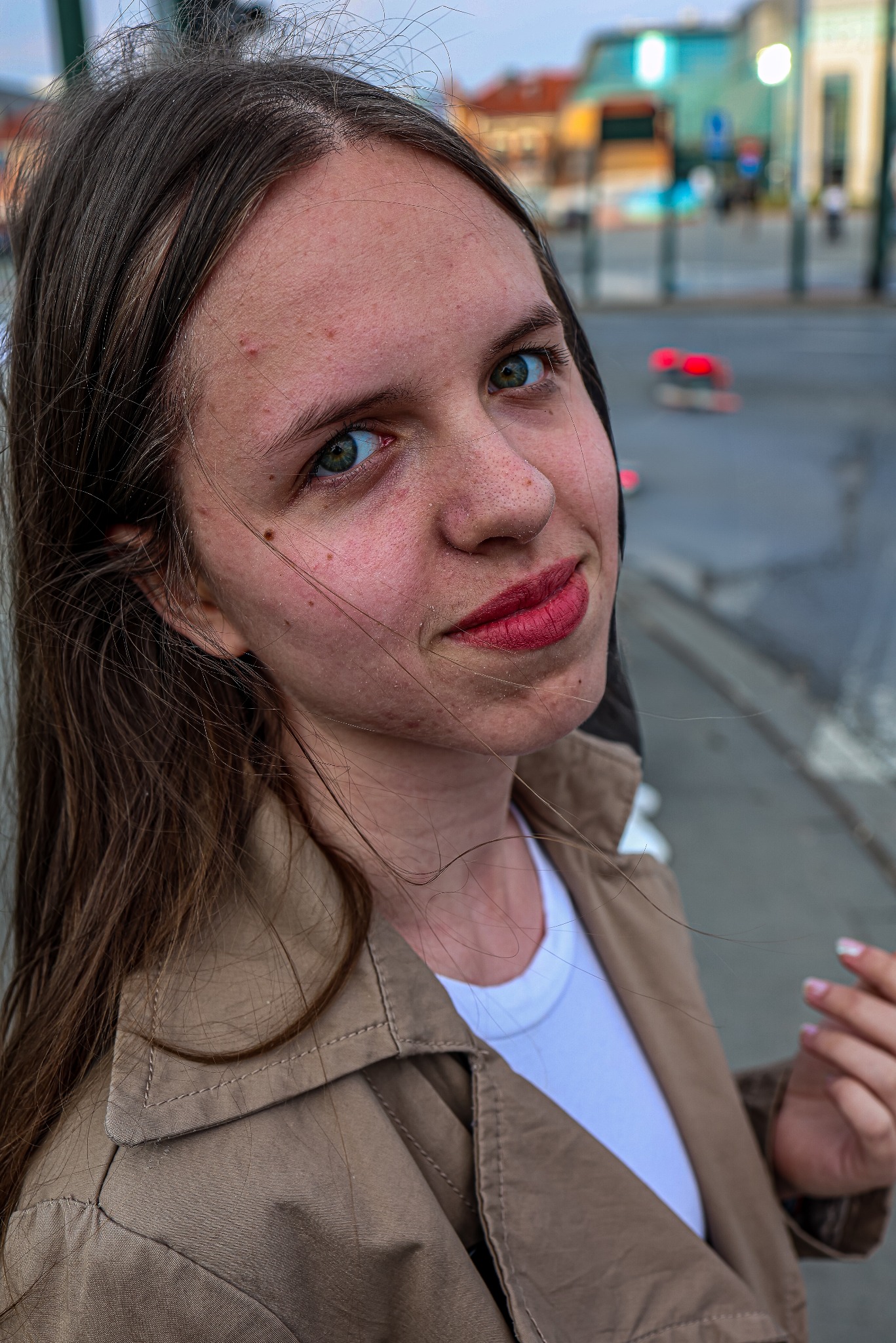 Portret młodej kobiety w beżowym płaszczu na tle miejskiej ulicy. Uwagę przyciągają jej zielone oczy i delikatny uśmiech. Widoczne detale skóry i włosów.