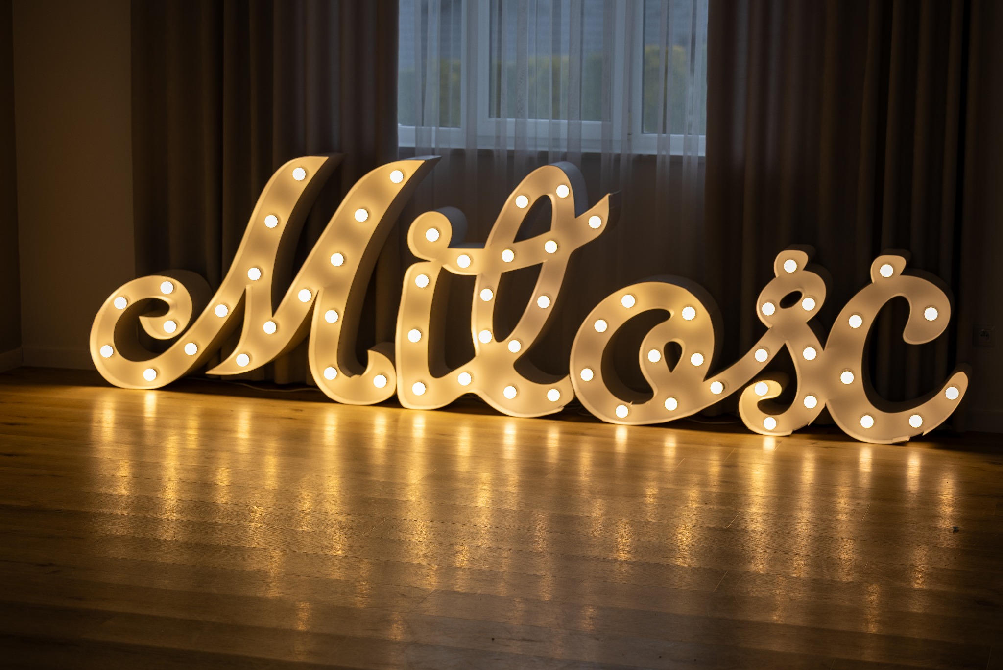 Świecący napis 'Miłość' z żarówkami na drewnianej podłodze, tło stanowią zasłony i okno. Dekoracja na sesję narzeczeńską lub ślubną, ciepłe światło.