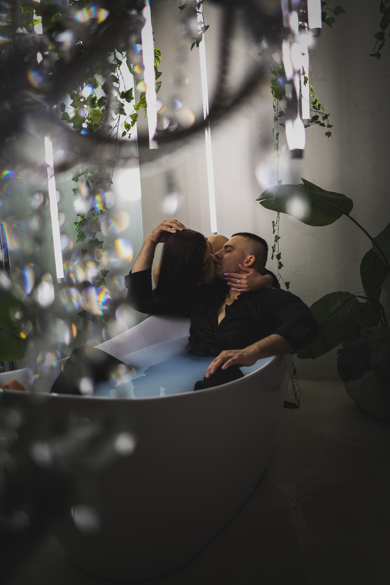 Zakochana para w czarnych koszulach, całująca się w wannie wypełnionej mleczną wodą, w otoczeniu roślin i dekoracji, w intymnej atmosferze.