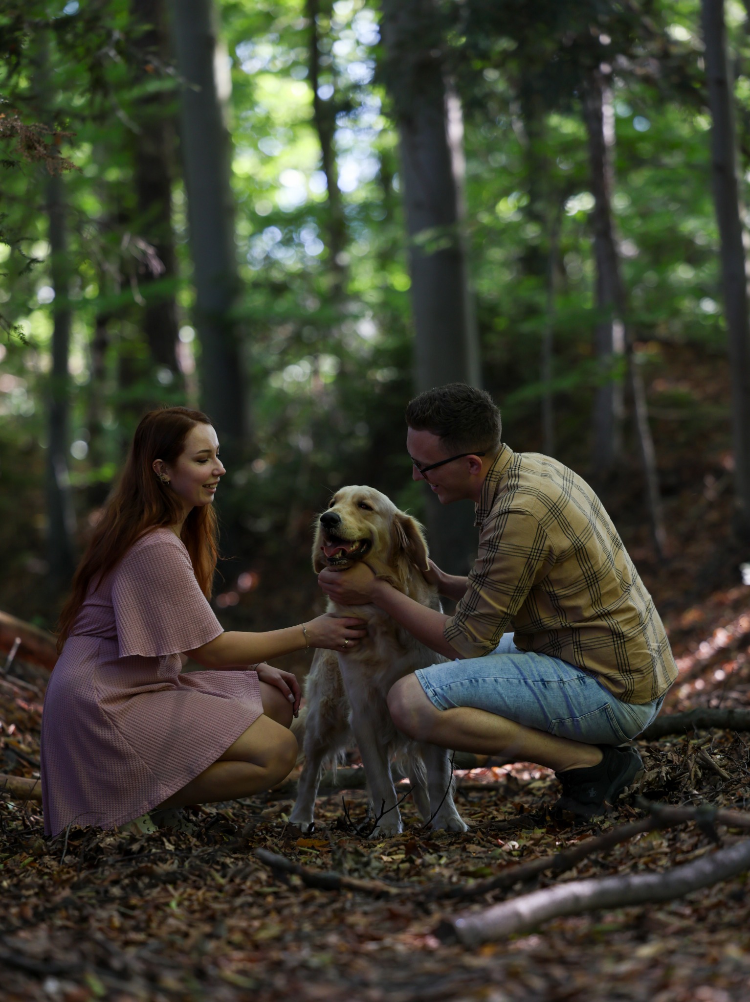 Para w lesie głaszcze golden retrievera. Kobieta w sukience i mężczyzna w koszuli w kratę kucają obok psa na tle drzew i opadłych liści.