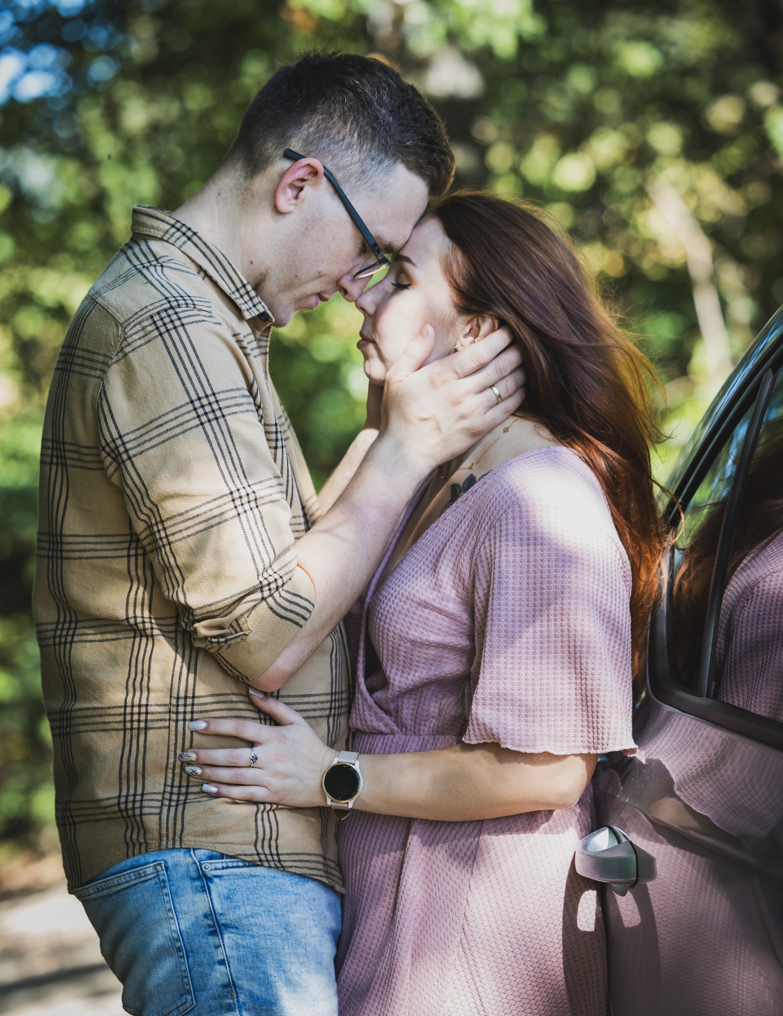 Zakochana para oparta o samochód, dotykająca się czołami na tle zieleni. Mężczyzna w kraciastej koszuli i jeansach, kobieta w różowej sukience. Romantyczna sesja.