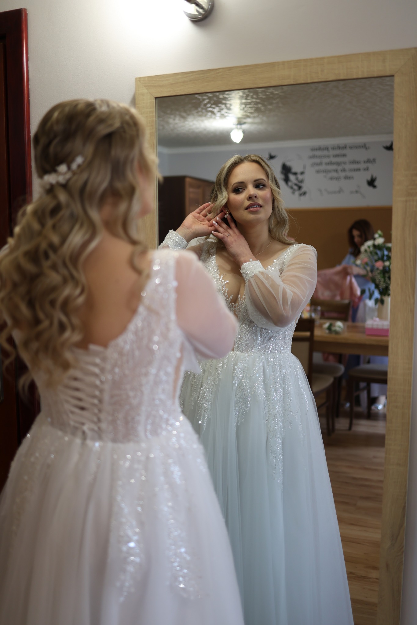 Panna młoda w białej sukni z koronkowym gorsetem i tiulowym dołem, poprawia kolczyk przed lustrem. W tle widoczna osoba i kwiaty.