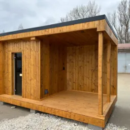 Nowoczesna, drewniana sauna zewnętrzna z przeszklonymi drzwiami i małym tarasem, umieszczona na betonowych bloczkach. Widok z zewnątrz, w tle budynek gospodarczy.