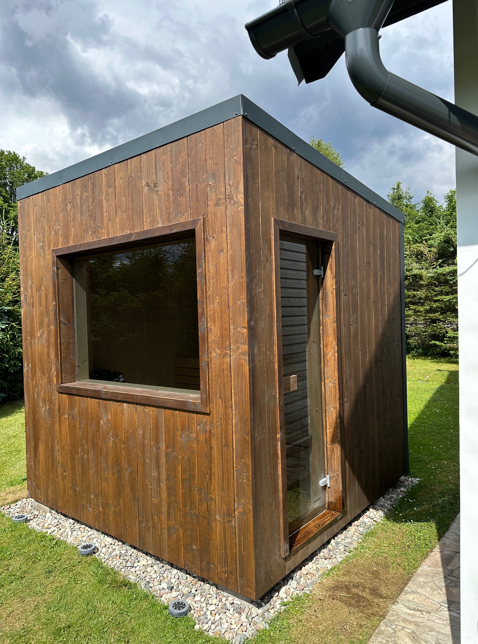 Nowoczesna, drewniana sauna ogrodowa z oknem i przeszklonymi drzwiami, otoczona kamieniami i trawnikiem w słoneczny dzień.