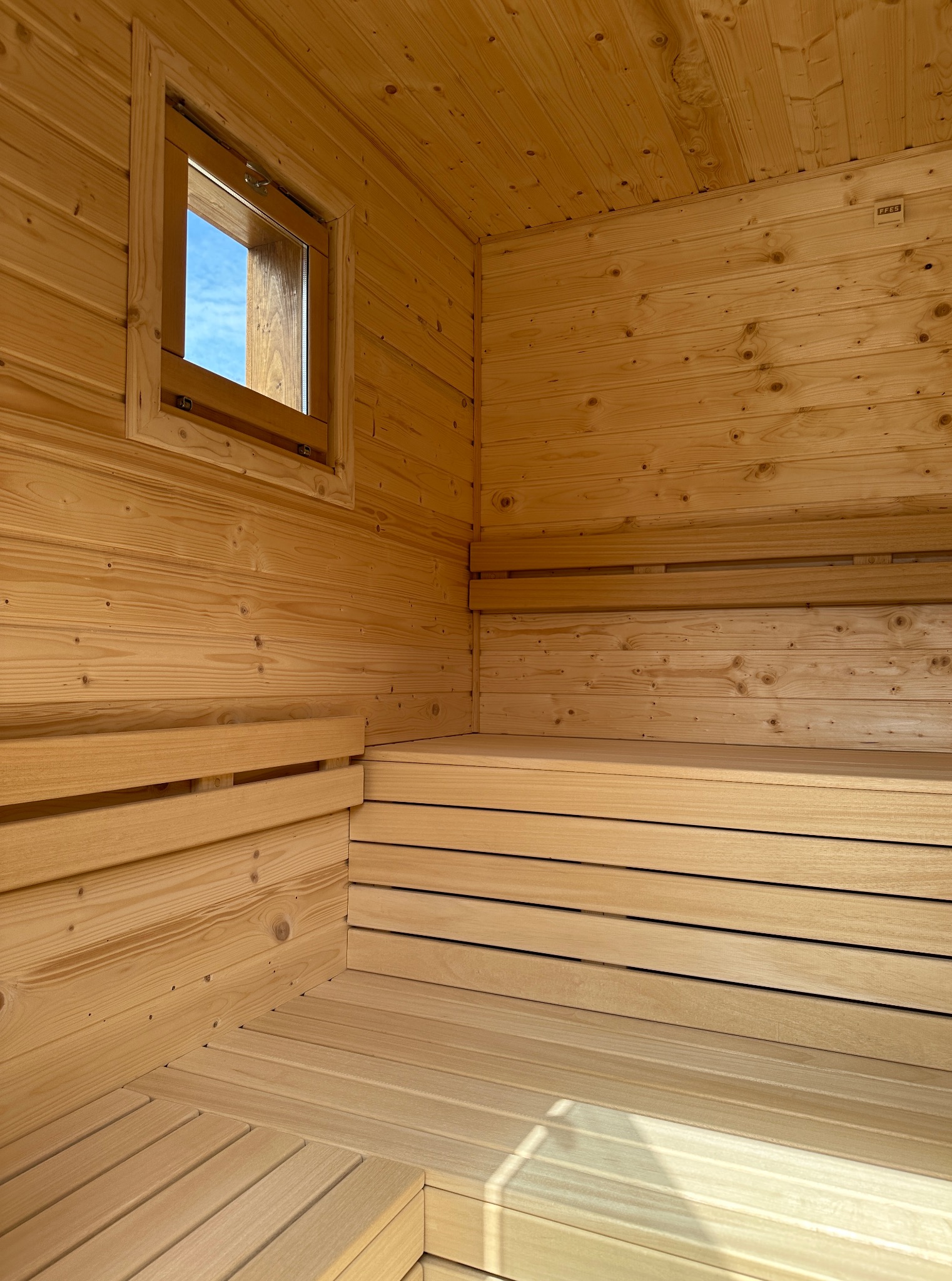 Jasne, drewniane wnętrze sauny z ławkami i oknem, przez które widać błękitne niebo. Widoczne naturalne usłojenie drewna i konstrukcja sauny.