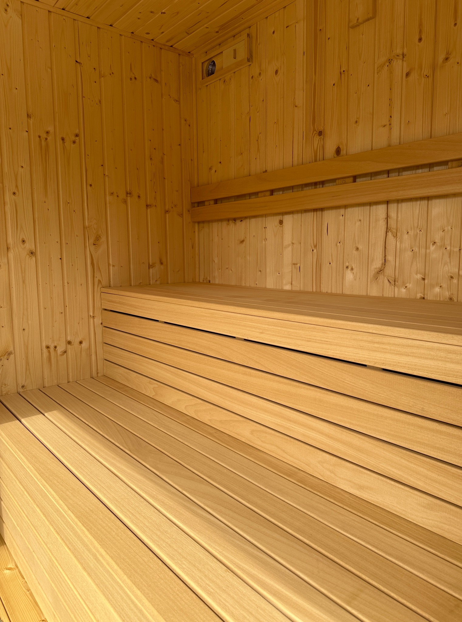 Jasne drewno w saunie: ławy i boazeria tworzą przytulne wnętrze. Widoczny wentylator w górnym rogu. Minimalistyczny design, ciepłe oświetlenie.