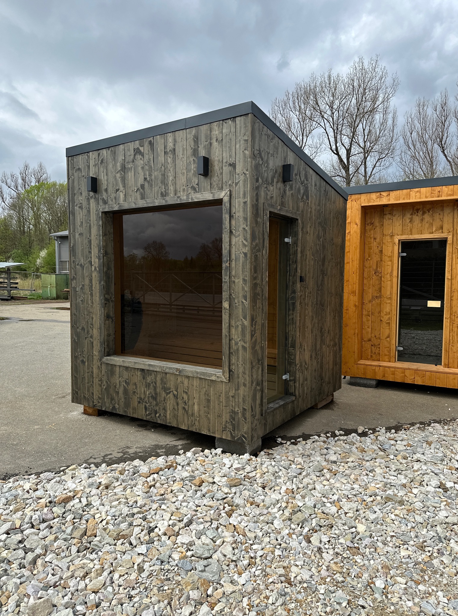 Nowoczesna, drewniana sauna zewnętrzna w odcieniach szarości, z dużym oknem i przeszklonymi drzwiami, ustawiona na żwirowym podłożu. Obok widoczna sauna w naturalnym kolorze drewna.