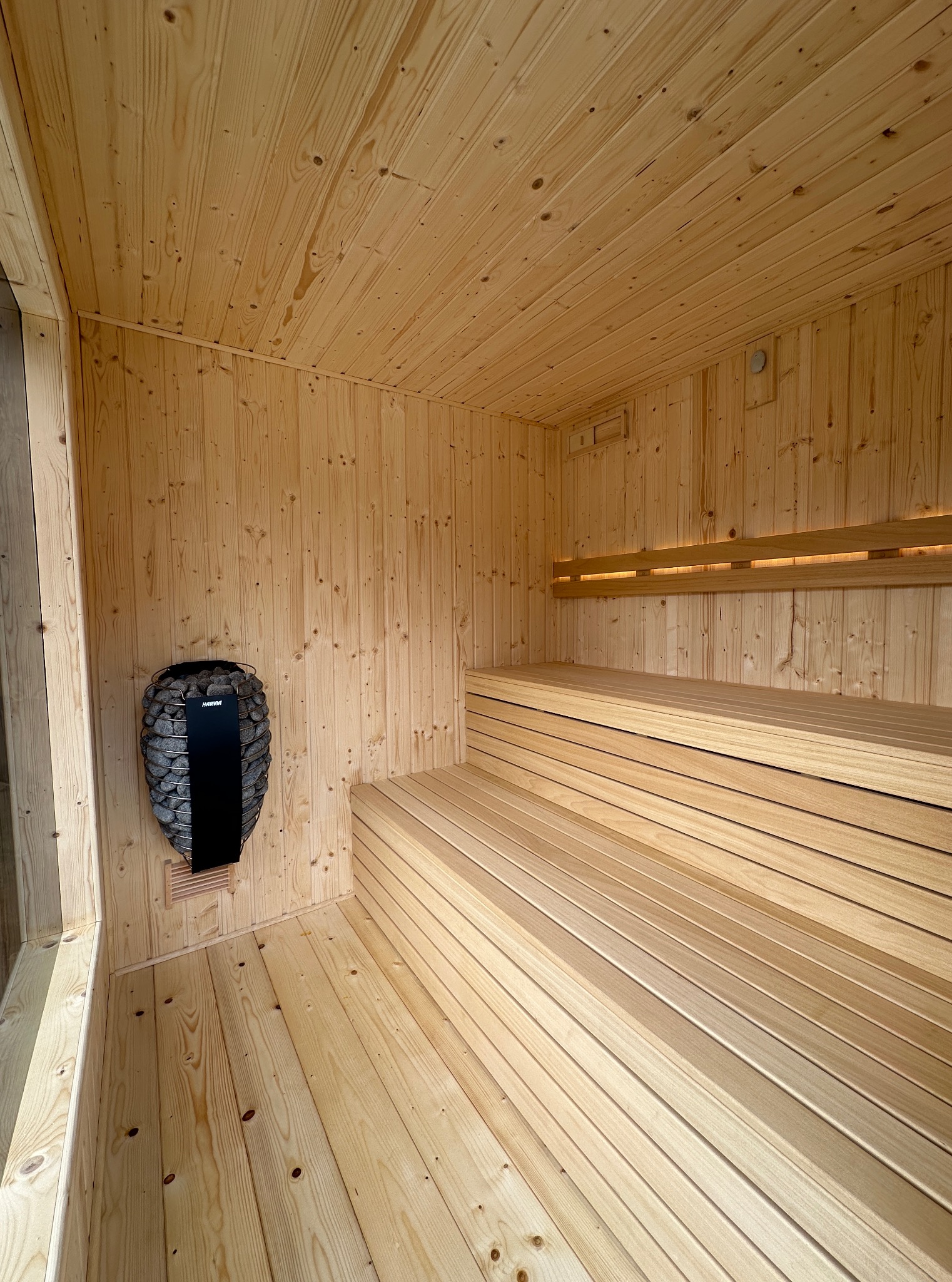Wnętrze sauny wykończone jasnym drewnem, z piecem Harvia na ścianie i trzypoziomowymi ławkami. Widoczne detale wykończenia i oświetlenie.