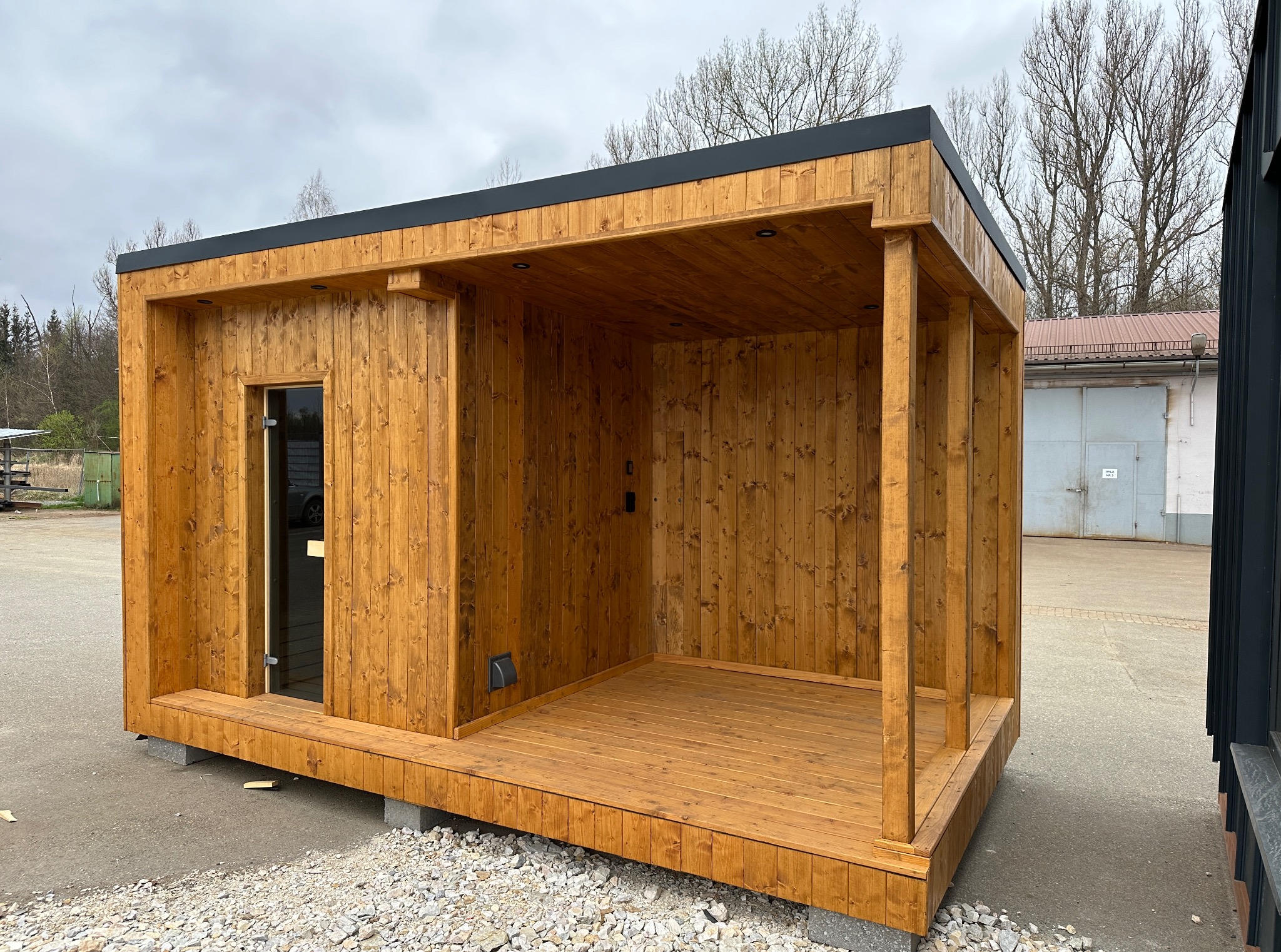 Nowoczesna, drewniana sauna zewnętrzna z przeszklonymi drzwiami i małym tarasem, umieszczona na betonowych bloczkach. Widok z zewnątrz, w tle budynek gospodarczy.