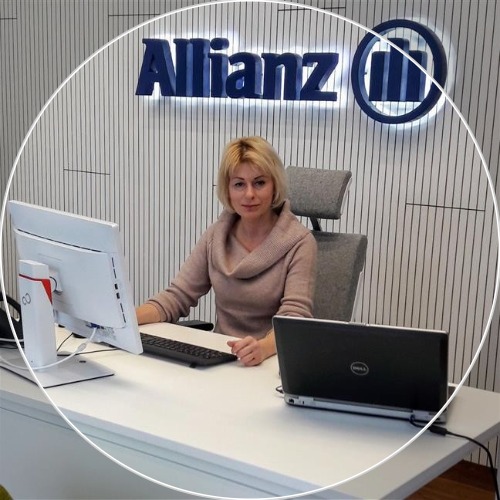 Kobieta w biurze Allianz siedzi przy biurku z komputerami. W tle logo firmy na ścianie z pionowymi paskami. Jasne oświetlenie biurowe.