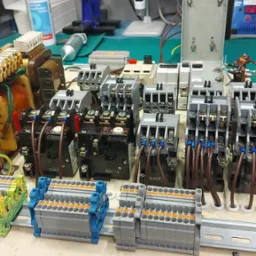 Panel sterowniczy z przekaźnikami i złączkami, widoczne okablowanie i elementy elektroniczne na stole warsztatowym z narzędziami w tle.