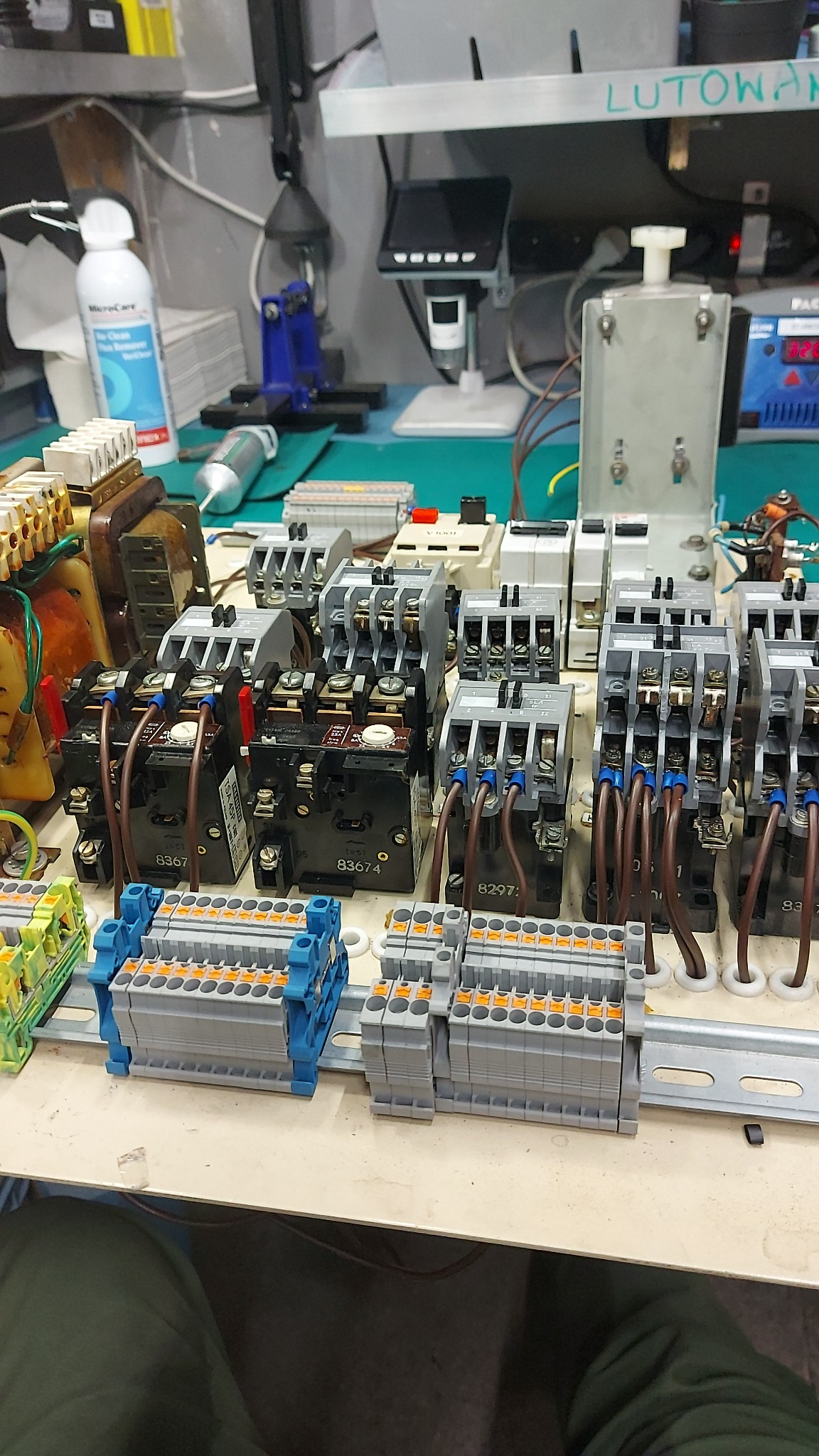 Panel sterowniczy z przekaźnikami i złączkami, widoczne okablowanie i elementy elektroniczne na stole warsztatowym z narzędziami w tle.