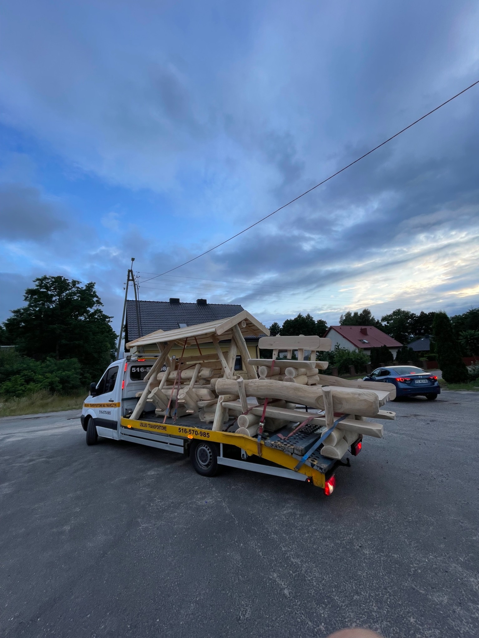 Laweta przewozi drewniany plac zabaw na tle domów i pochmurnego nieba. Widoczny numer kontaktowy na samochodzie. Usługa transportowa w toku.