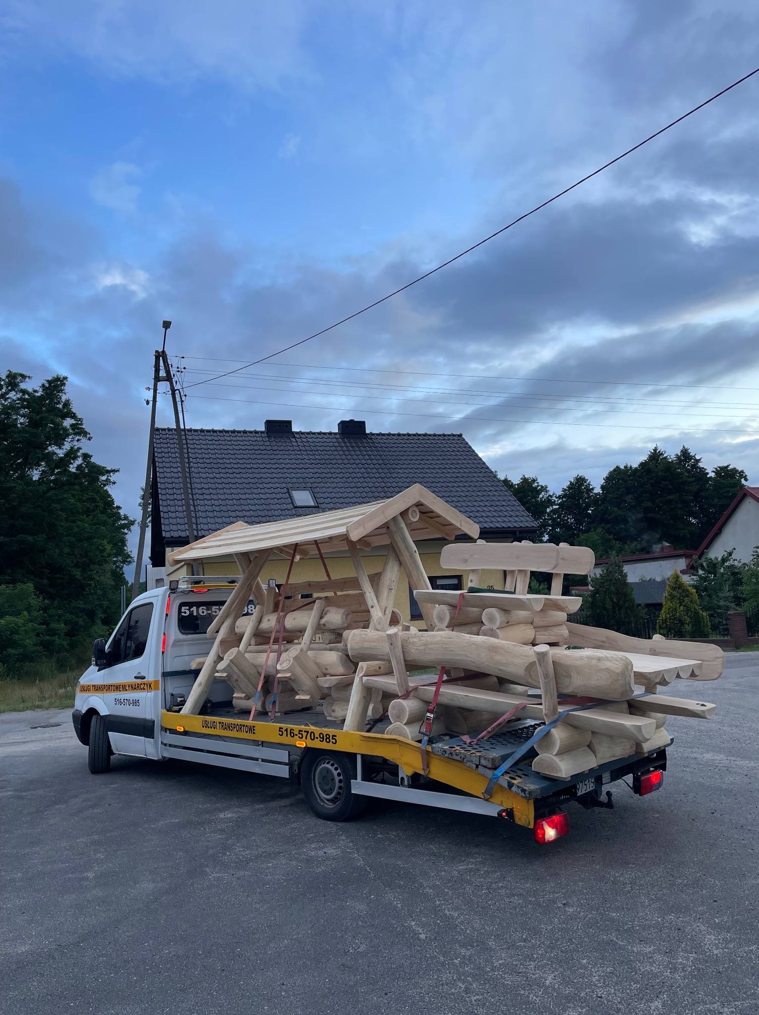 Laweta przewozi drewniany plac zabaw z bali na tle domów i pochmurnego nieba. Na lawecie widoczne mocowania i pasy zabezpieczające ładunek. Usługi transportowe Młynarczyk.