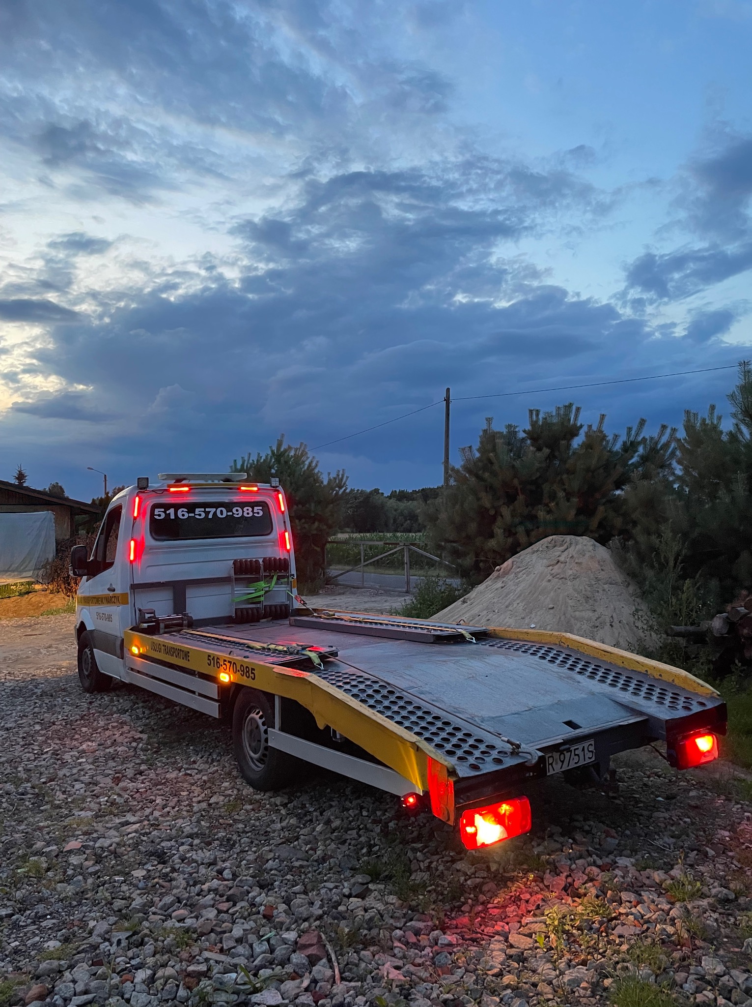 Laweta z podświetlonym numerem telefonu na tle pochmurnego nieba i wiejskiego krajobrazu. Widoczne światła stopu i tablica rejestracyjna.