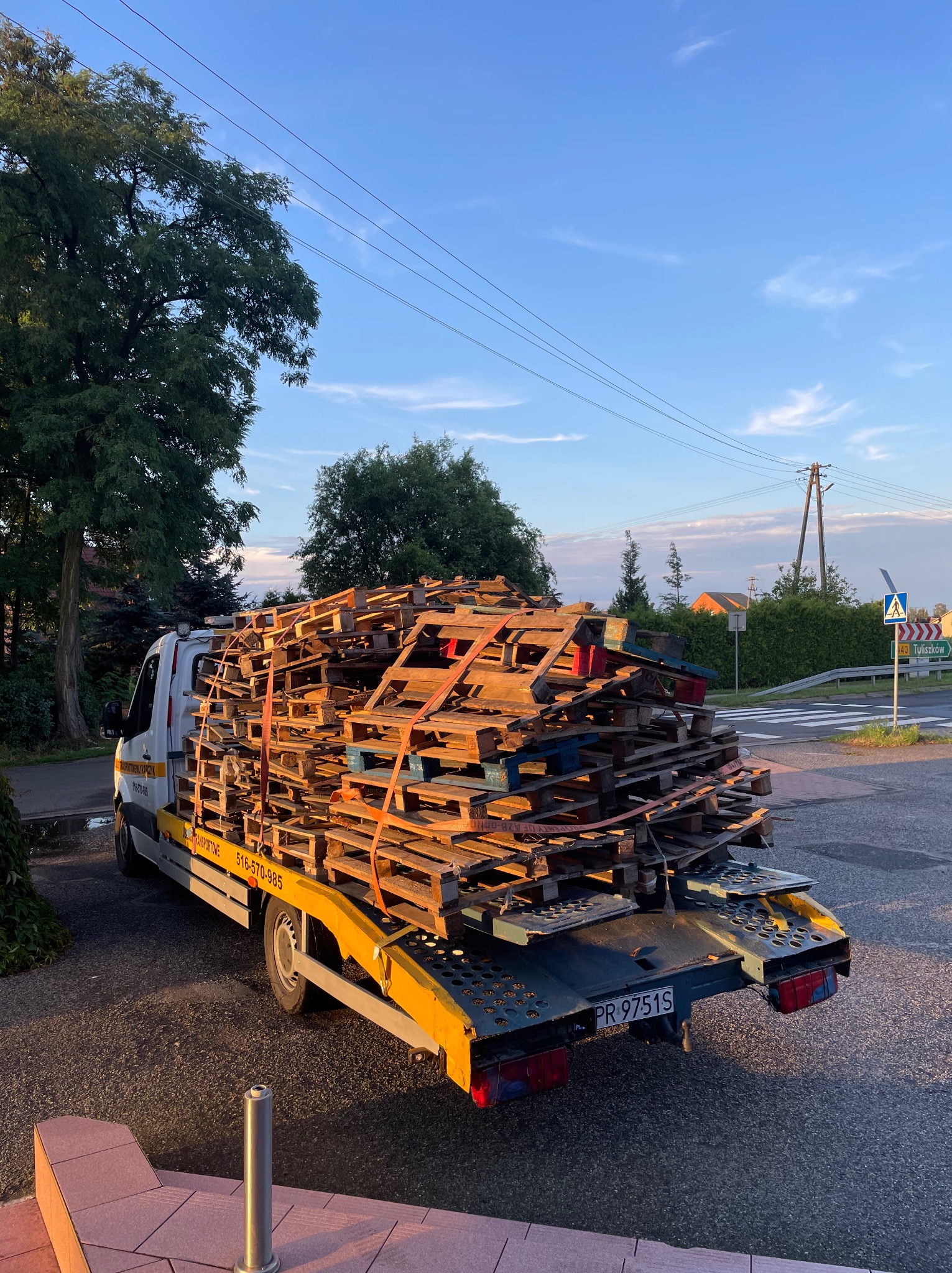 Laweta z platformą przewozi duży ładunek drewnianych palet, zabezpieczonych pasami, na tle skrzyżowania i błękitnego nieba o zmierzchu.