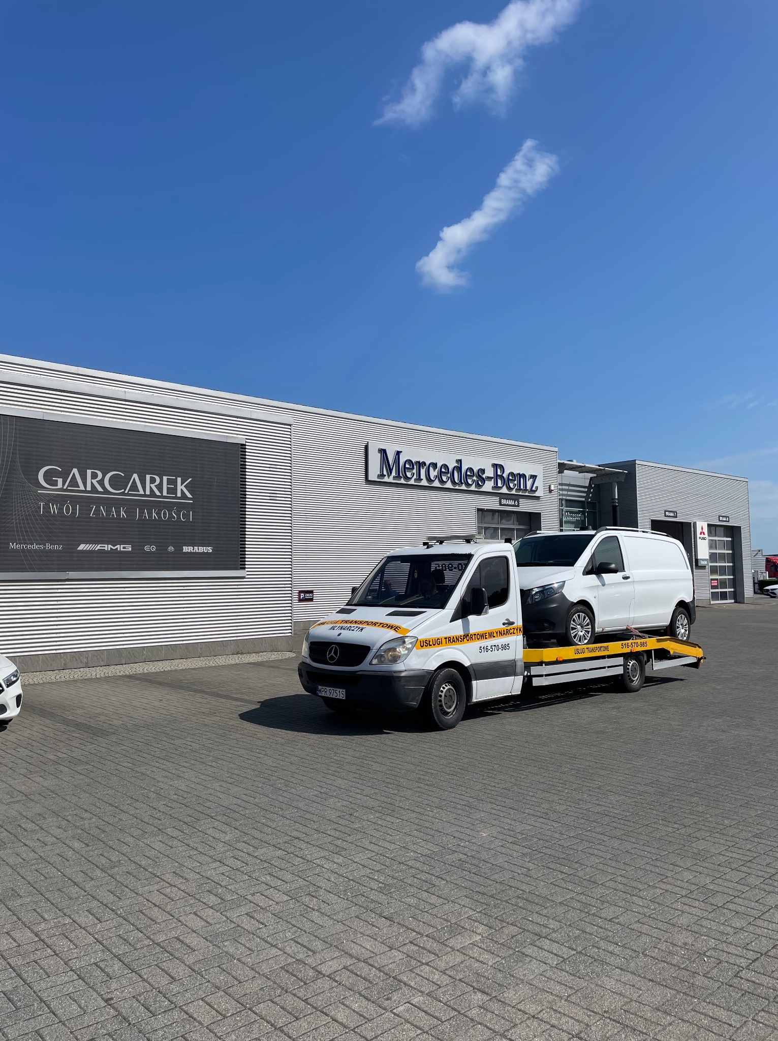 Laweta z białym busem na tle salonu Mercedes-Benz. Żółta platforma lawety z napisem „Usługi Transportowe Młynarczyk”. Błękitne niebo w tle.