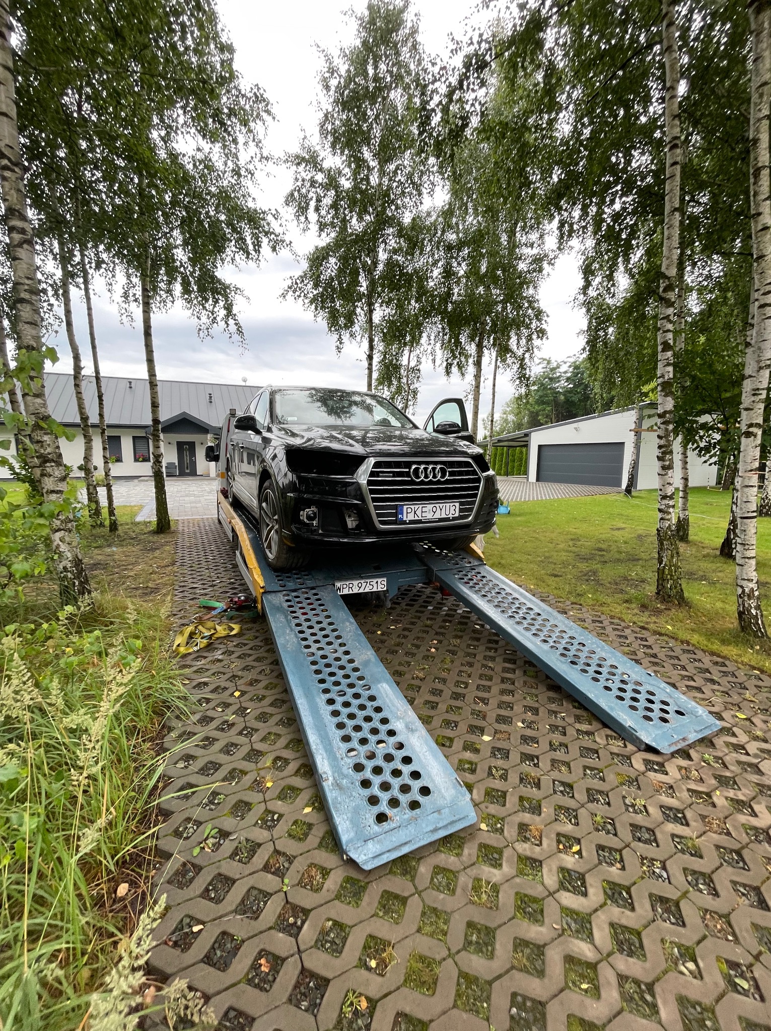 Czarny samochód Audi na lawecie, z otwartymi drzwiami, na tle domu i drzew. Tablica rejestracyjna PKE 9YU3. Rampy w kolorze niebieskim.