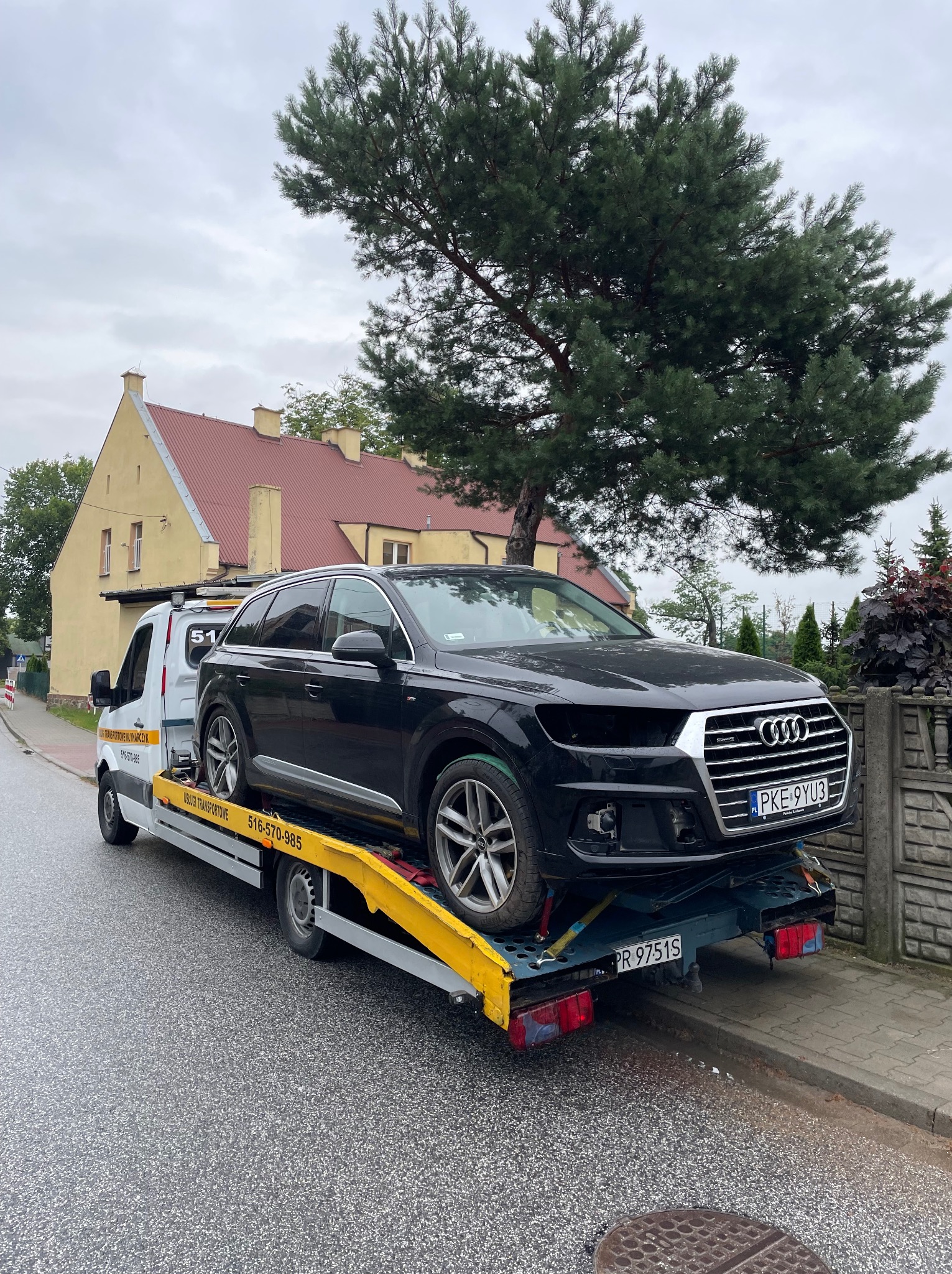 Czarny SUV Audi Q7 na żółtej lawecie na tle ulicy i budynku z czerwoną dachówką. Widoczny numer rejestracyjny pojazdu. Pochmurne niebo.